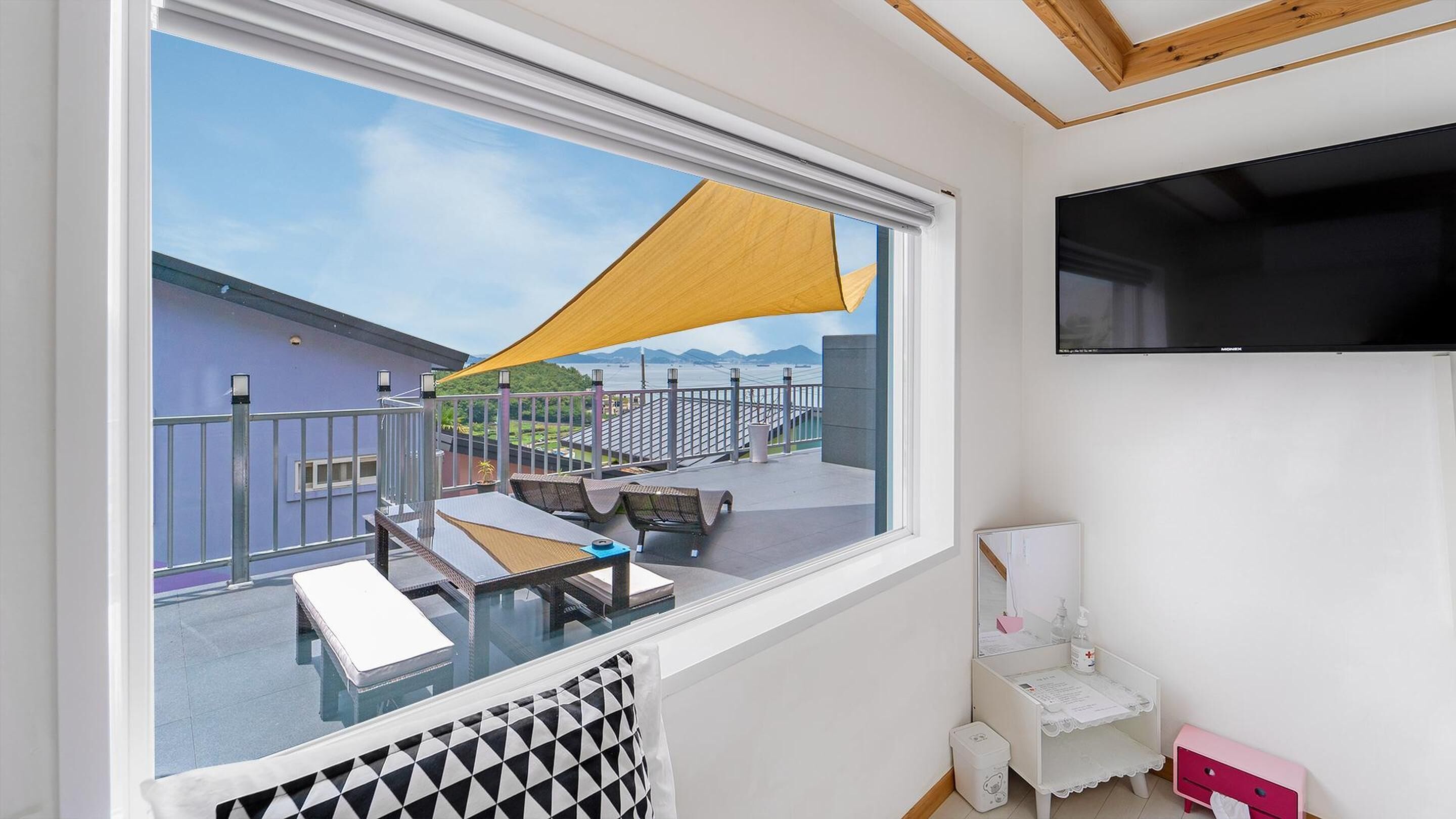 Ein Balkon mit Blick auf ein Gebäude und Berge, ein an der Wand befestigter Fernseher und ein kleines, weißes Schränkchen mit Spiegel.