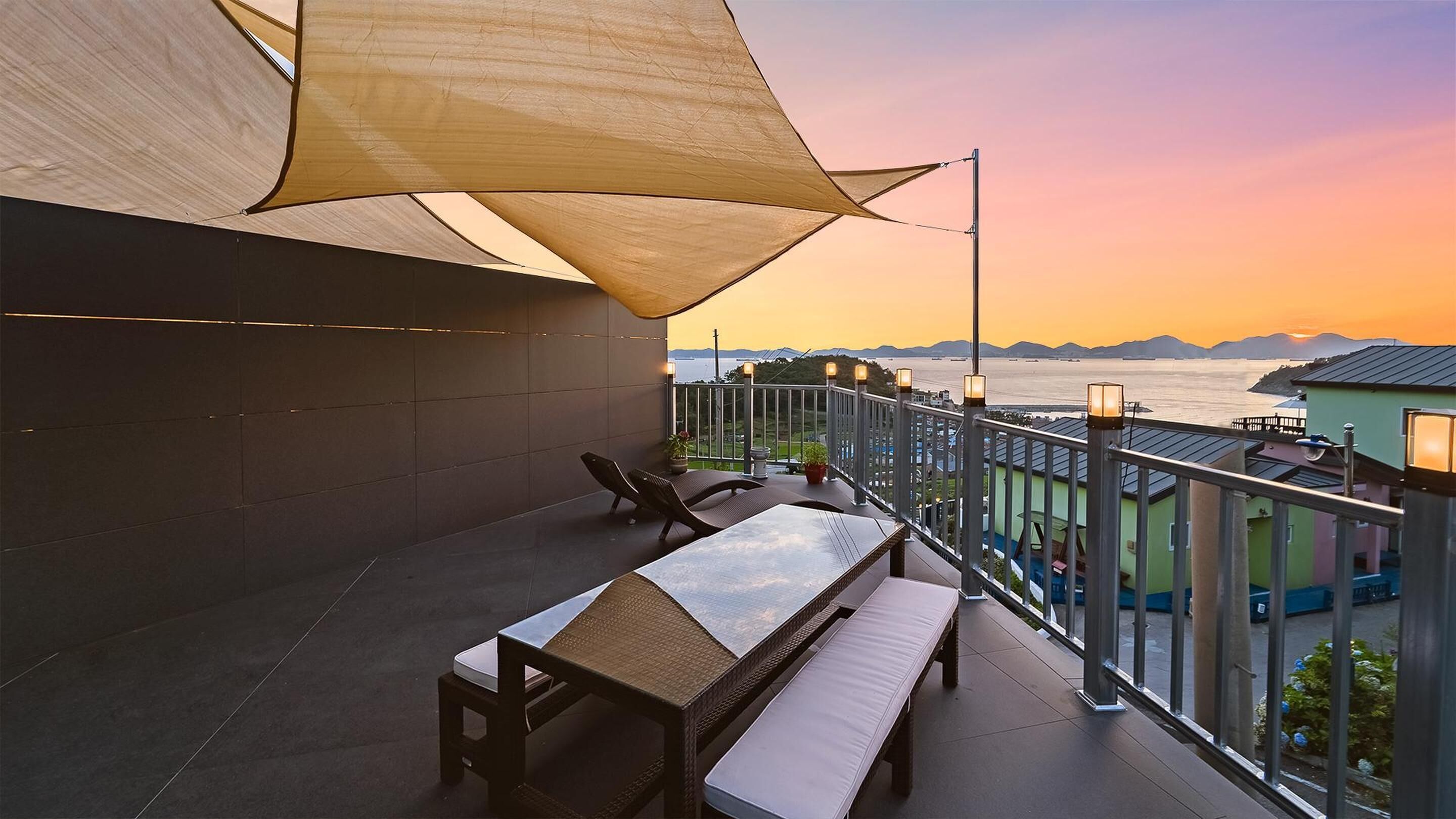 Ein Balkon mit Meerblick bei Sonnenuntergang, ausgestattet mit Tisch und Stühlen sowie einem großen Sonnensegel.