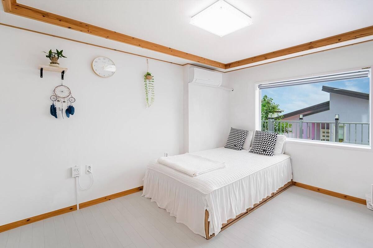 A bedroom with a bed, a window, a wall-mounted air conditioner, a shelf with a plant, and a dreamcatcher.
