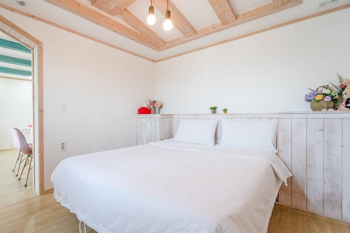 A bedroom with a wooden ceiling, a bed, a bench, a chair, and a small table with a vase of flowers.