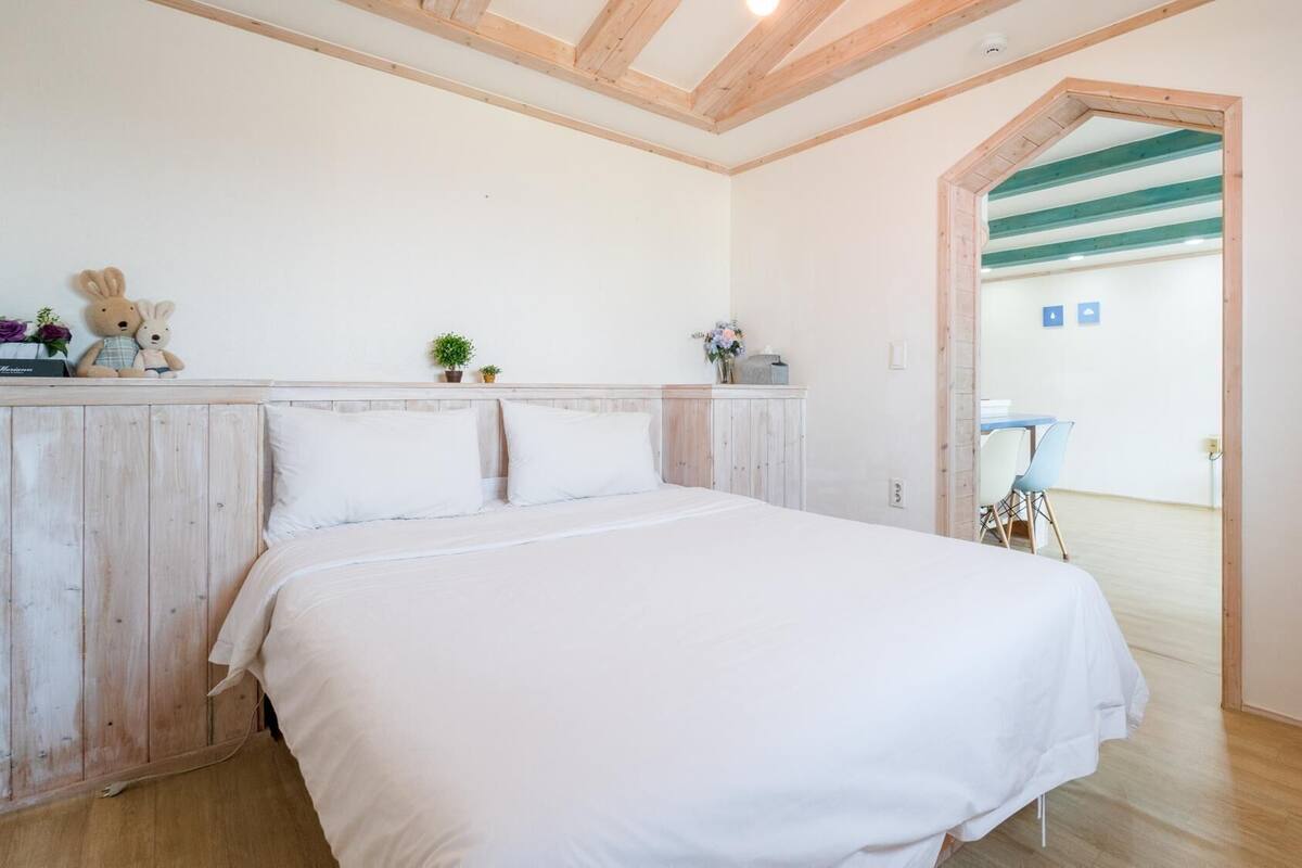 A bedroom with a wooden headboard, a bed with white bedding, a small desk with a chair, and a door leading to another room.