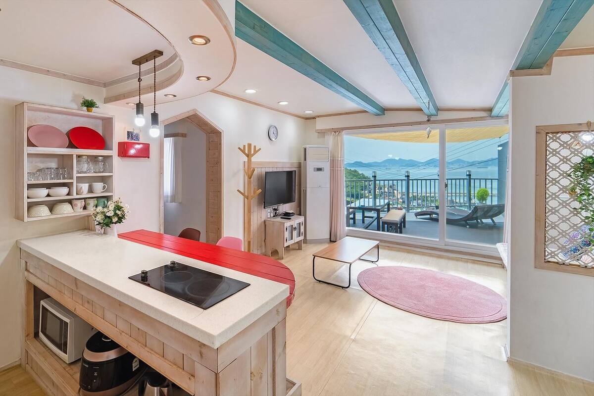 A modern kitchen with a built-in stove, a dining area with a red table, and a balcony with a view of the mountains.
