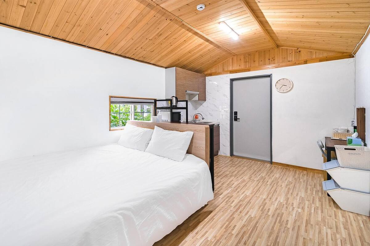 A modern bedroom with a wooden ceiling, a bed with white bedding, a small kitchenette, and a view of greenery through a window.