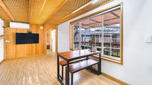 A room with wooden walls and ceiling, a large window with a view of a building, a flat-screen TV mounted on the wall, and a wooden table with benches. - Jeju Bijalim Pension (Jeju City)