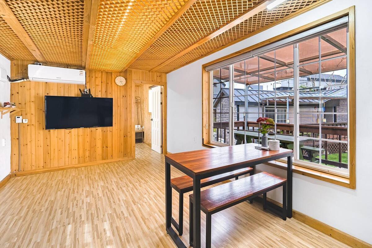 A room with wooden walls and ceiling, a large window with a view of a building, a flat-screen TV mounted on the wall, and a wooden table with benches.