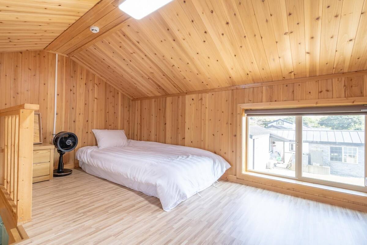 A wooden room with a bed, a window, and a fan.
