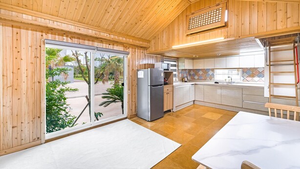 A modern kitchen with wooden walls, a large sliding glass door, and a view of a tropical outdoor area. - Jeju Bijalim Pension (Jeju City)