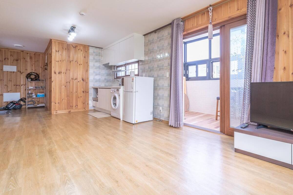 A spacious living area with a kitchen, a washing machine, a television, and a sliding glass door leading to a balcony.
