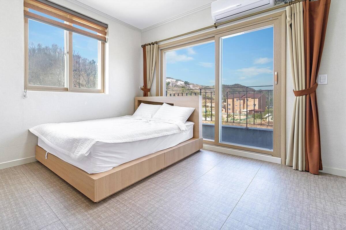 Un dormitorio con cama, ventanas amplias y vista a edificios.