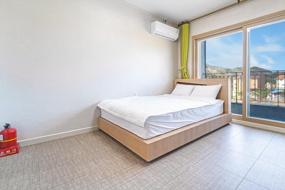 Un dormitorio moderno con cama, extintor y ventana grande con vistas a edificios y colinas.