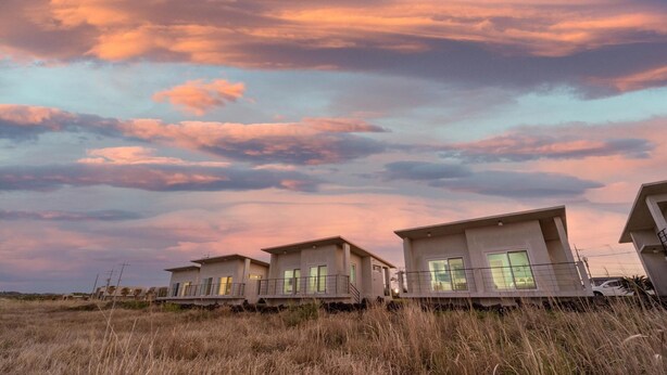 A row of modern, single-story houses with large windows and flat roofs, situated in a grassy field with a dramatic sky in the background. - Jeju Byeoleuldameunbaramarae Pension (Jeju City)