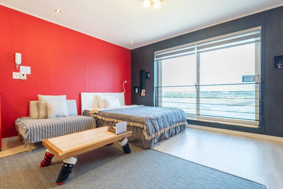 A modern hotel room with a red accent wall, a large window with a view, a kitchenette, and a bed with a grey duvet.