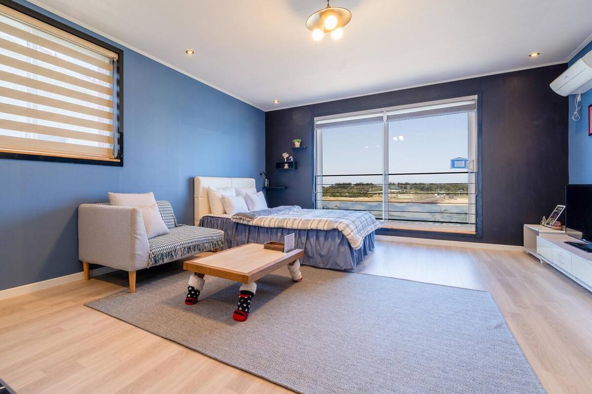 A modern bedroom with a large window, a sofa, a bed, and a small table.