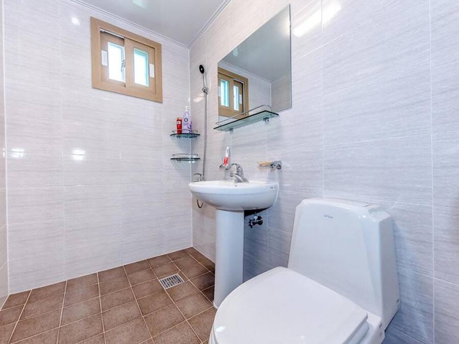 A bathroom with white tiles, a white toilet, a sink with a mirror above it, and a window with a wooden frame.