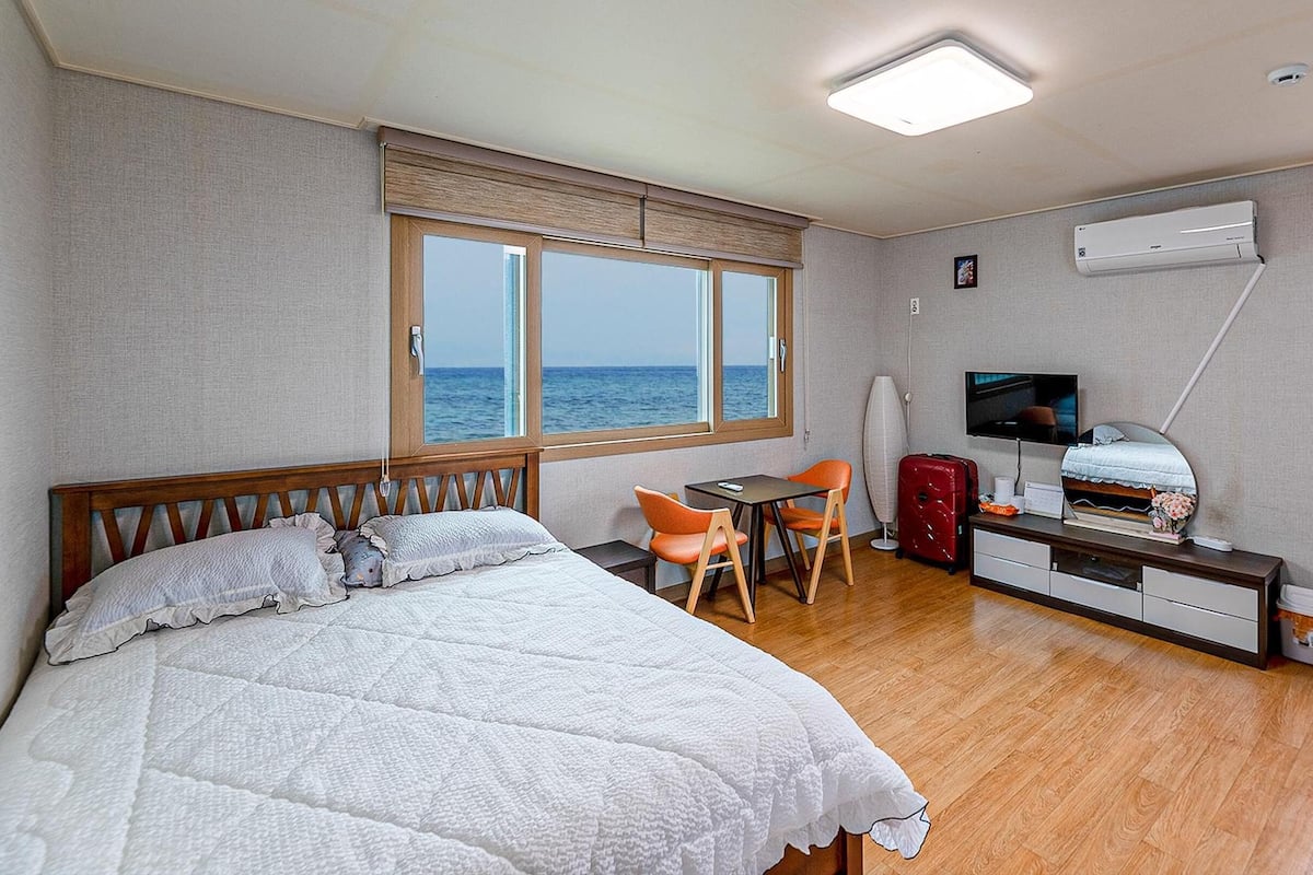 A hotel room with a bed, a desk with a chair, a TV, and a window with a view of the sea.