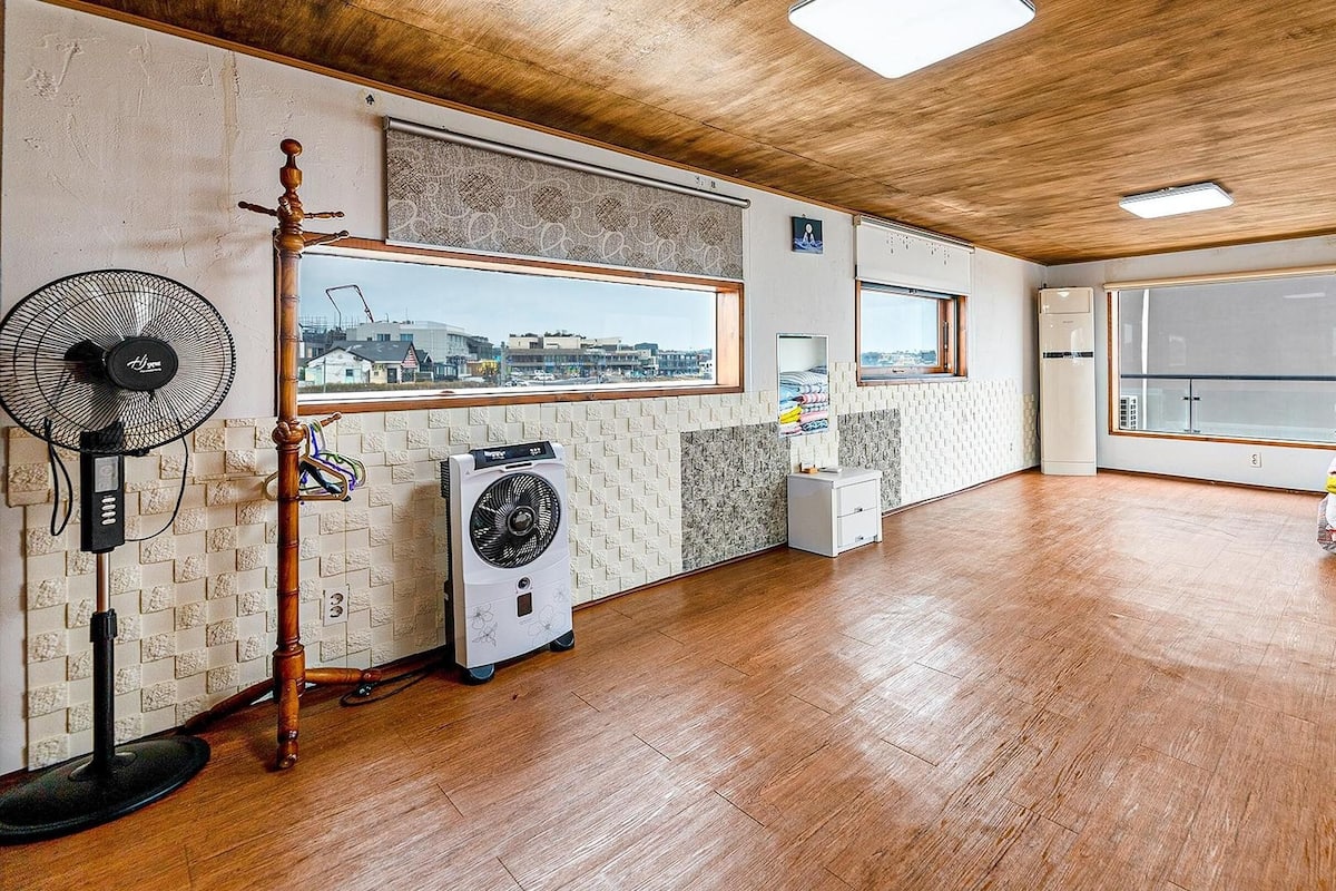 A room with a wooden floor, a standing fan, a portable air conditioner, and a window with a city view.
