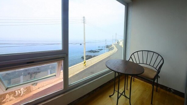 A room with a large window overlooking a coastal road and sea, a small round table, and a metal chair. - Jeju Syagaluibada Pension (Jeju City)