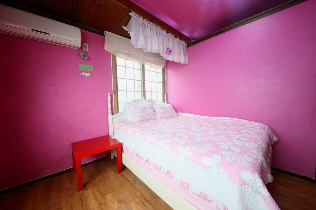 Un dormitorio con paredes rosas, suelo de madera, una cama con colcha estampada, un banco rojo y una ventana con cortinas blancas.