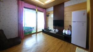 A compact kitchen with a refrigerator, a microwave, and a sink. There is a window with pink curtains and a view of greenery outside. - Jeju Syagaluibada Pension (Jeju City)