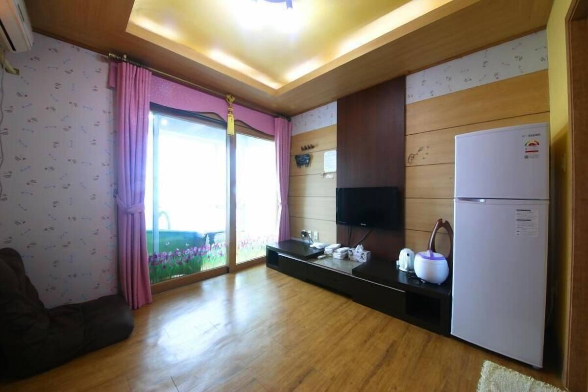Cocina compacta con refrigerador, microondas y lavamanos. Se ve una ventana con cortinas rosadas y vistas a la vegetación.