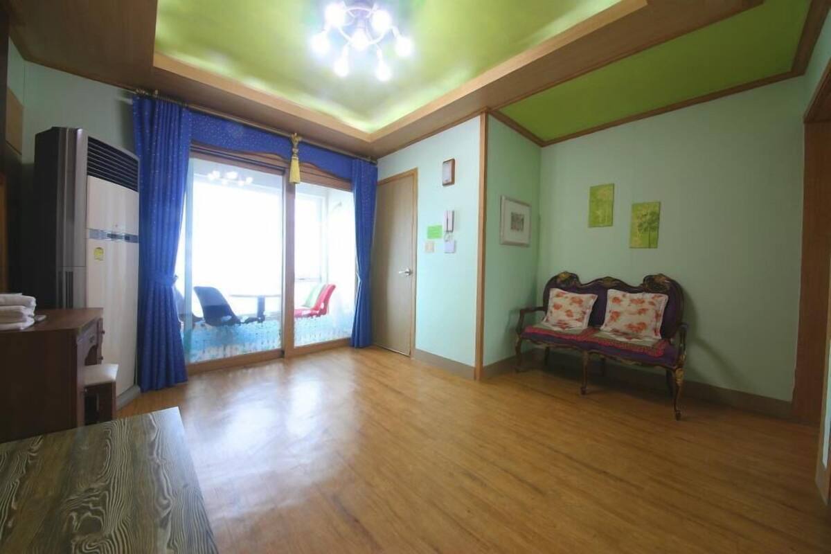 Una habitación amplia con piso de madera, un sofá con cojines estampados, un ventilador de techo y un ventanal corredizo que da a un balcón.