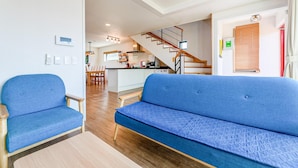 Un salon moderne avec un canapé et des fauteuils bleus, un escalier en bois et une cuisine en arrière-plan.