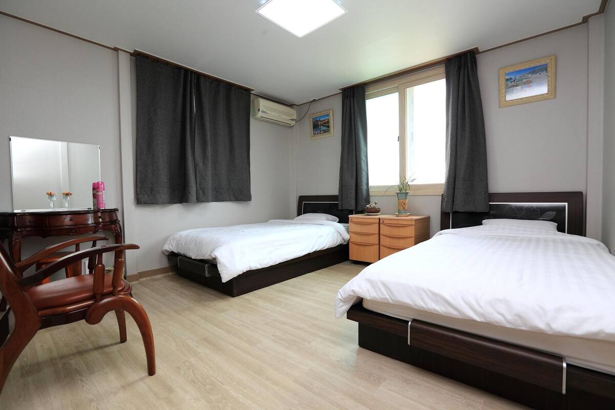 Une chambre d’hôtel avec deux lits simples, une chaise en bois, un bureau avec un miroir, une commode et une fenêtre avec des rideaux.