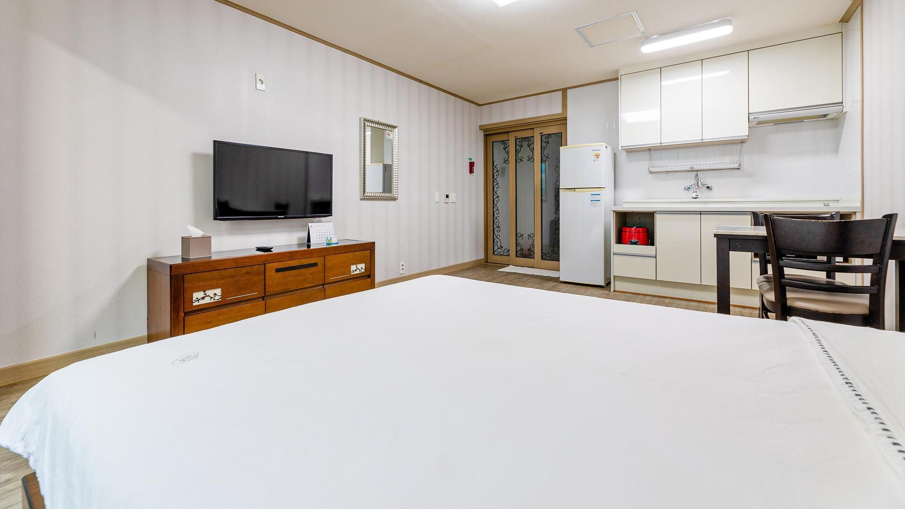 A hotel room with a large bed, a TV, a wooden dresser, a desk with a chair, a refrigerator, and a kitchen area with cabinets.
