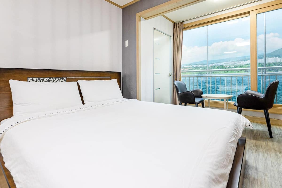 Une chambre d’hôtel avec un grand lit, une petite table, deux chaises et un balcon offrant une vue sur l’eau et le paysage urbain.