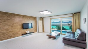 A modern living room with a flat-screen TV, a sofa, a coffee table, and a large sliding glass door leading to a balcony with a view of the sea and mountains. - Geoje Rockspa Pension (Geoje)