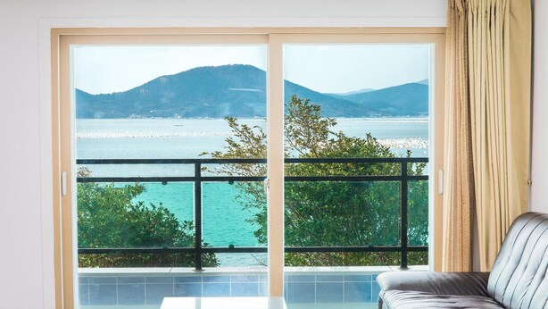 A room with a large sliding glass door leading to a balcony with a view of a body of water and hills. - Geoje Rockspa Pension (Geoje)