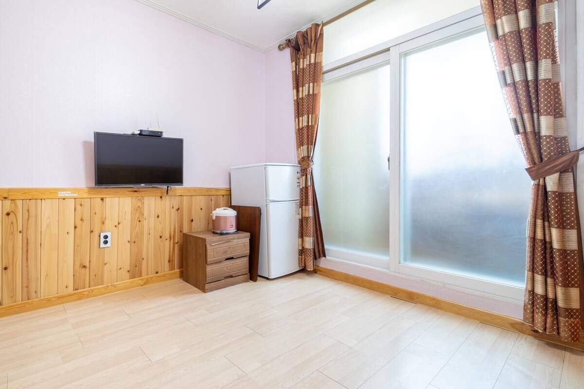 A room with a wooden floor, a television on a wooden shelf, a refrigerator, and a window with curtains.