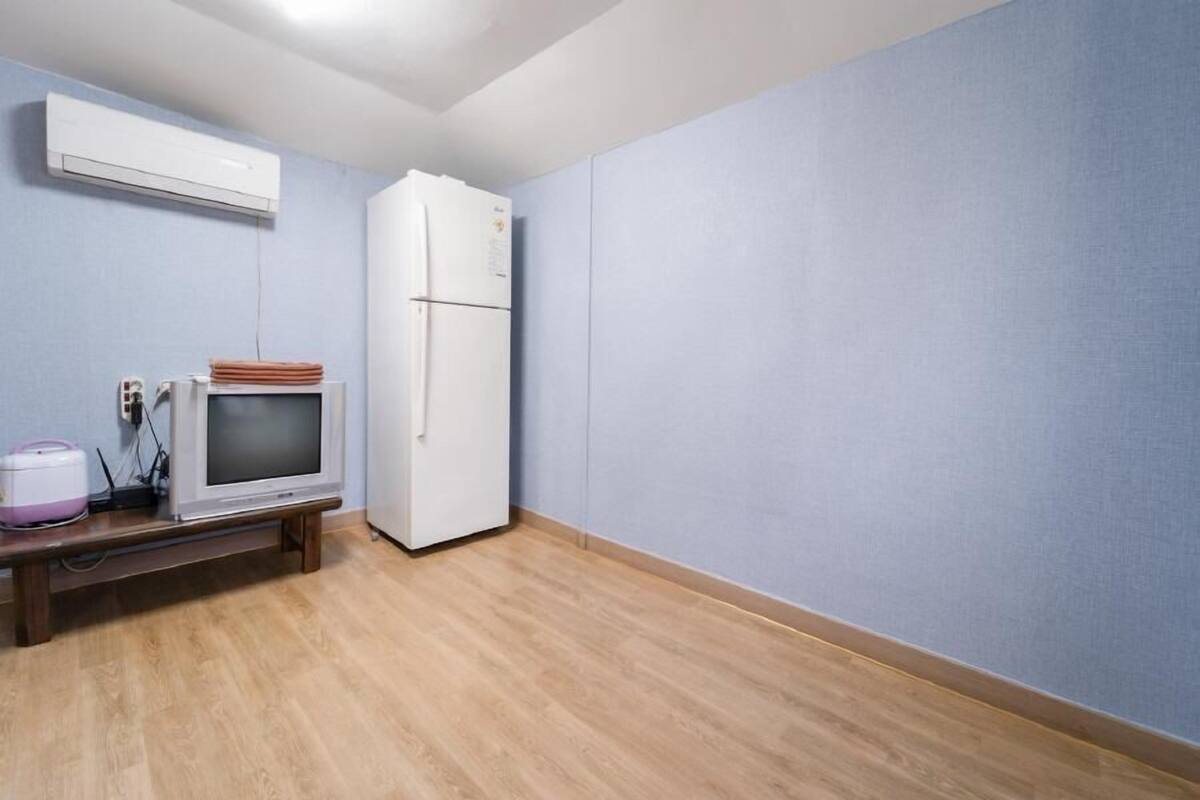 A room with a wooden floor, a white refrigerator, a flat-screen TV on a wooden stand, and a wall-mounted air conditioner.