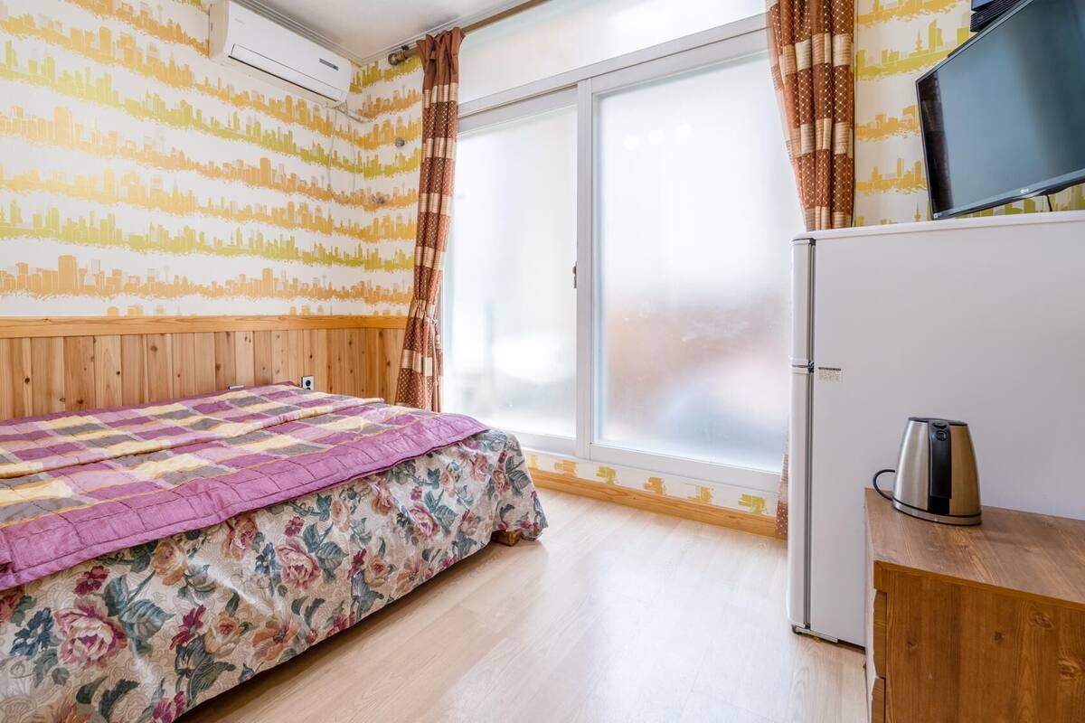 A bedroom with a bed, a wooden headboard, a window with curtains, a television, and a refrigerator.