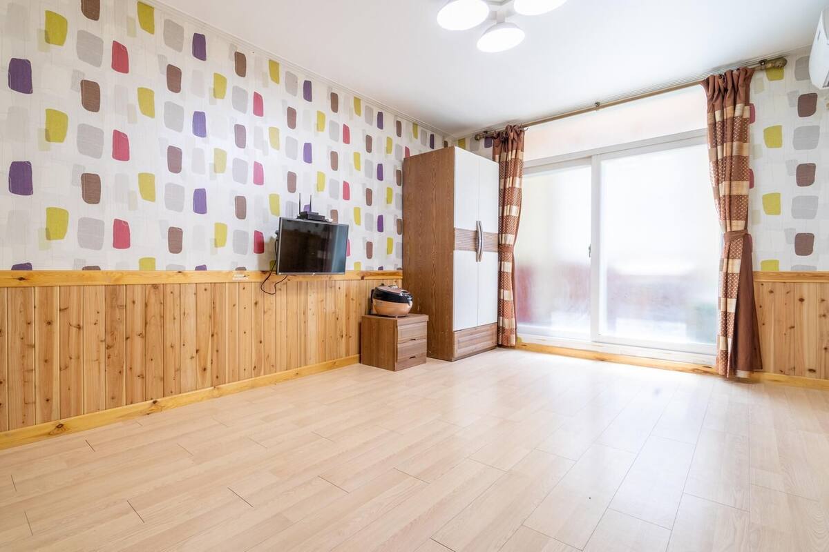 A spacious room with wooden paneling, a large window with curtains, and a television on a wooden stand.