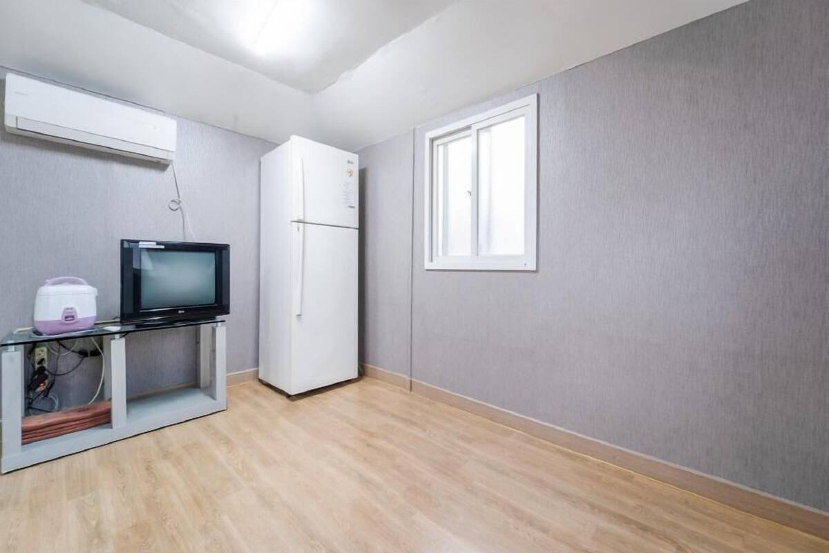 A small, modern room with a flat-screen TV on a stand, a white refrigerator, a window, and a wooden floor.