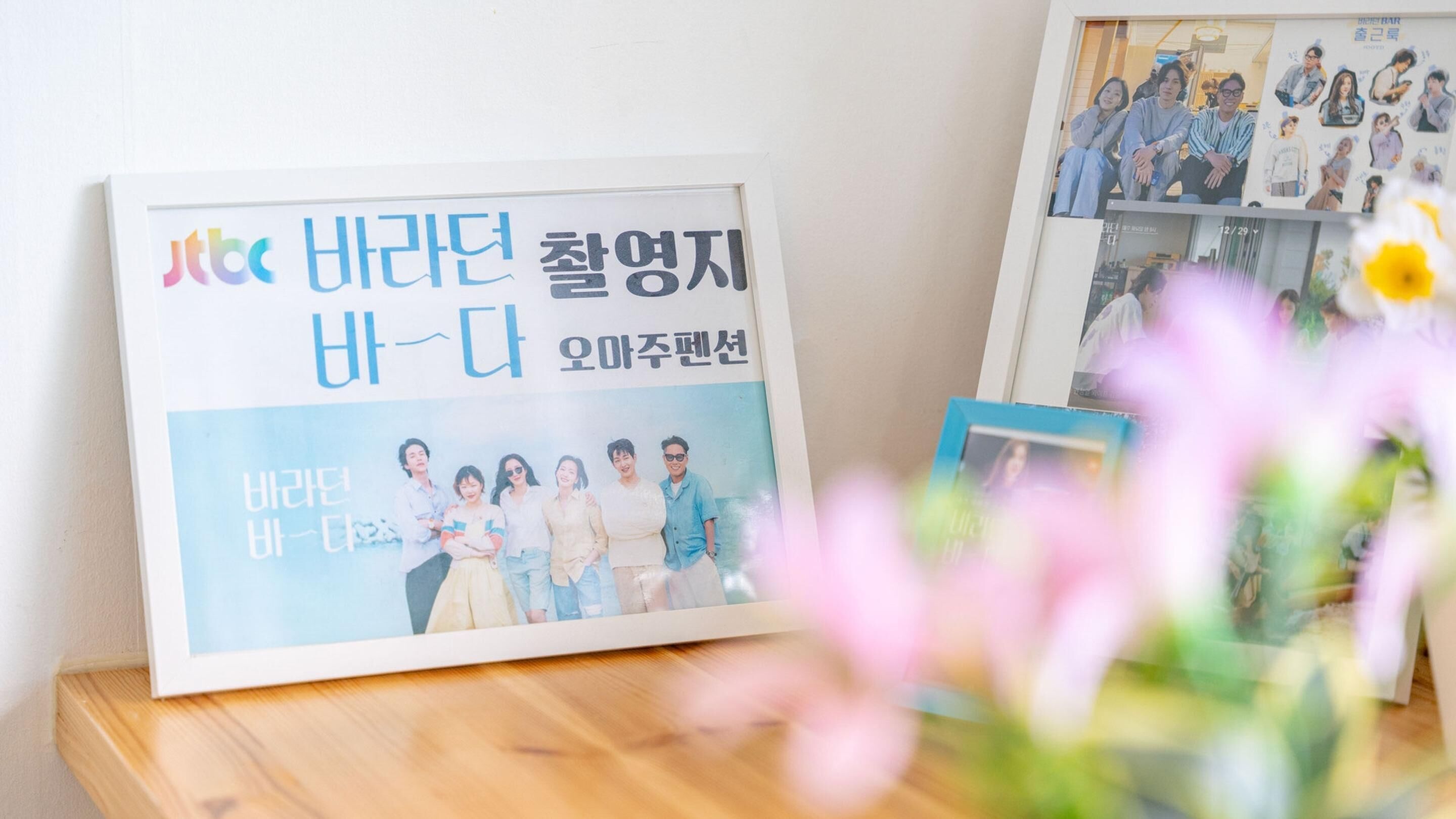 Ein gerahmtes Poster mit koreanischem Text und einem Gruppenfoto, platziert auf einer Holzfläche vor unscharfem Hintergrund.