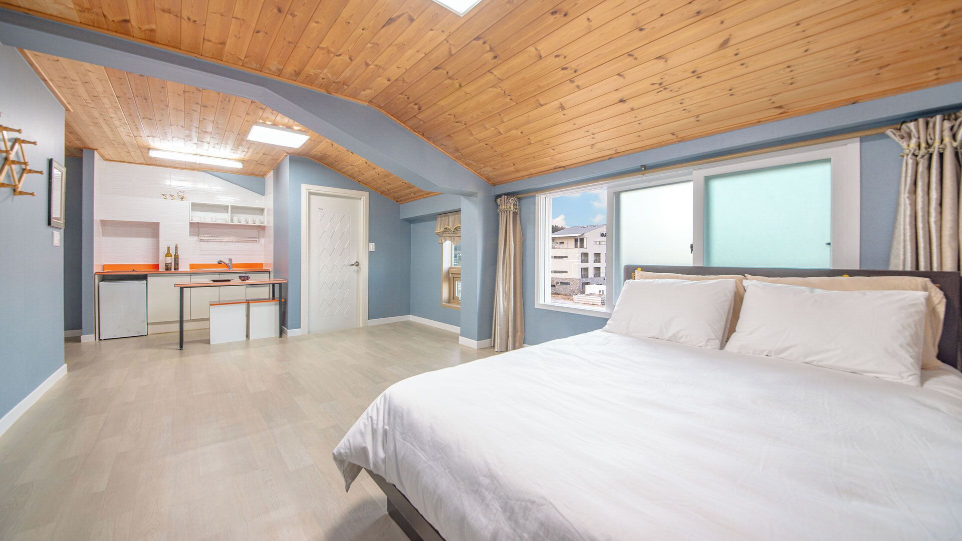 Quarto com uma cama grande, vista pela janela e uma área de cozinha ao fundo.