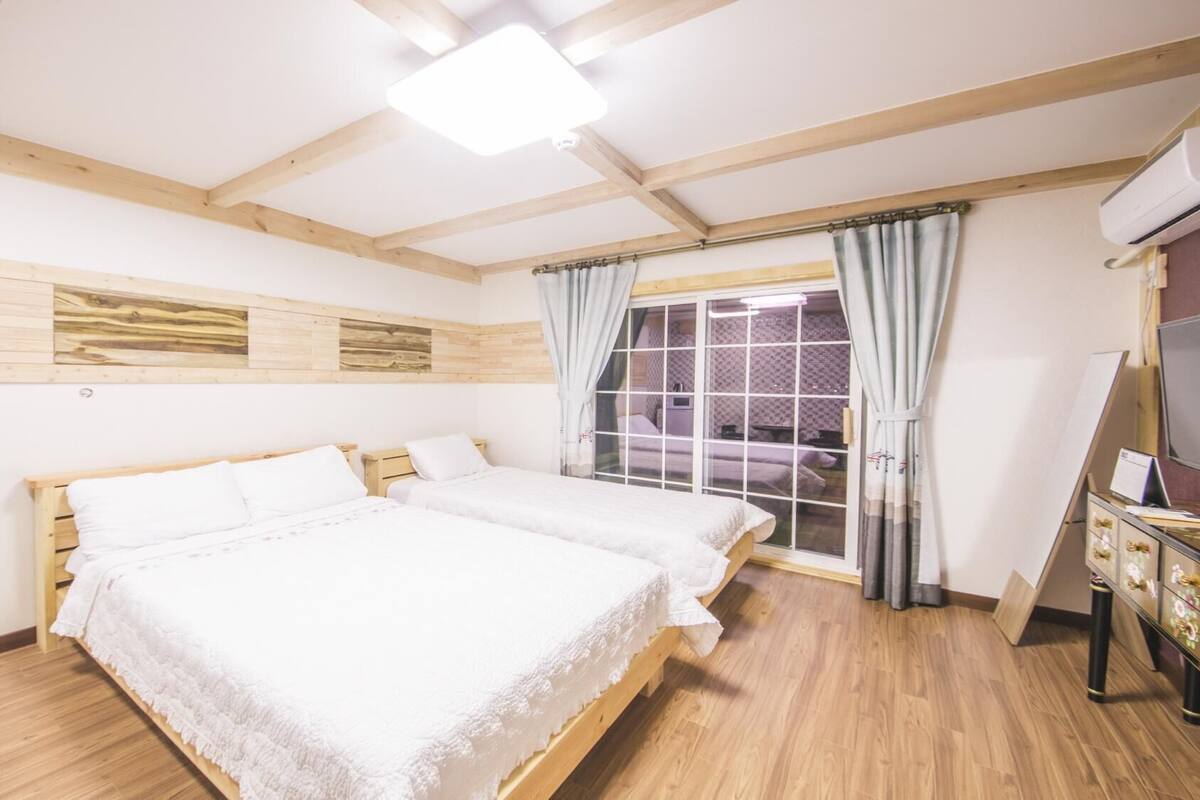 A room with two beds, wooden beams, a window with curtains, and an air conditioning unit.