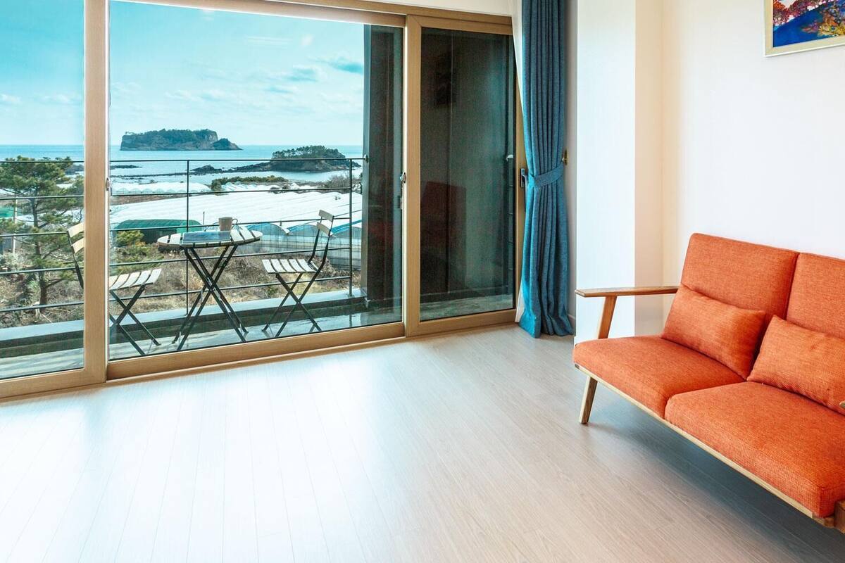 A room with a large sliding glass door leading to a balcony with a view of the sea and a small table with chairs.