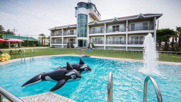 A swimming pool with inflatable orca figures, a fountain, and a multi-story building with balconies in the background. - Seogwipo Jejuhae-anhyuyang Pension (Seogwipo)