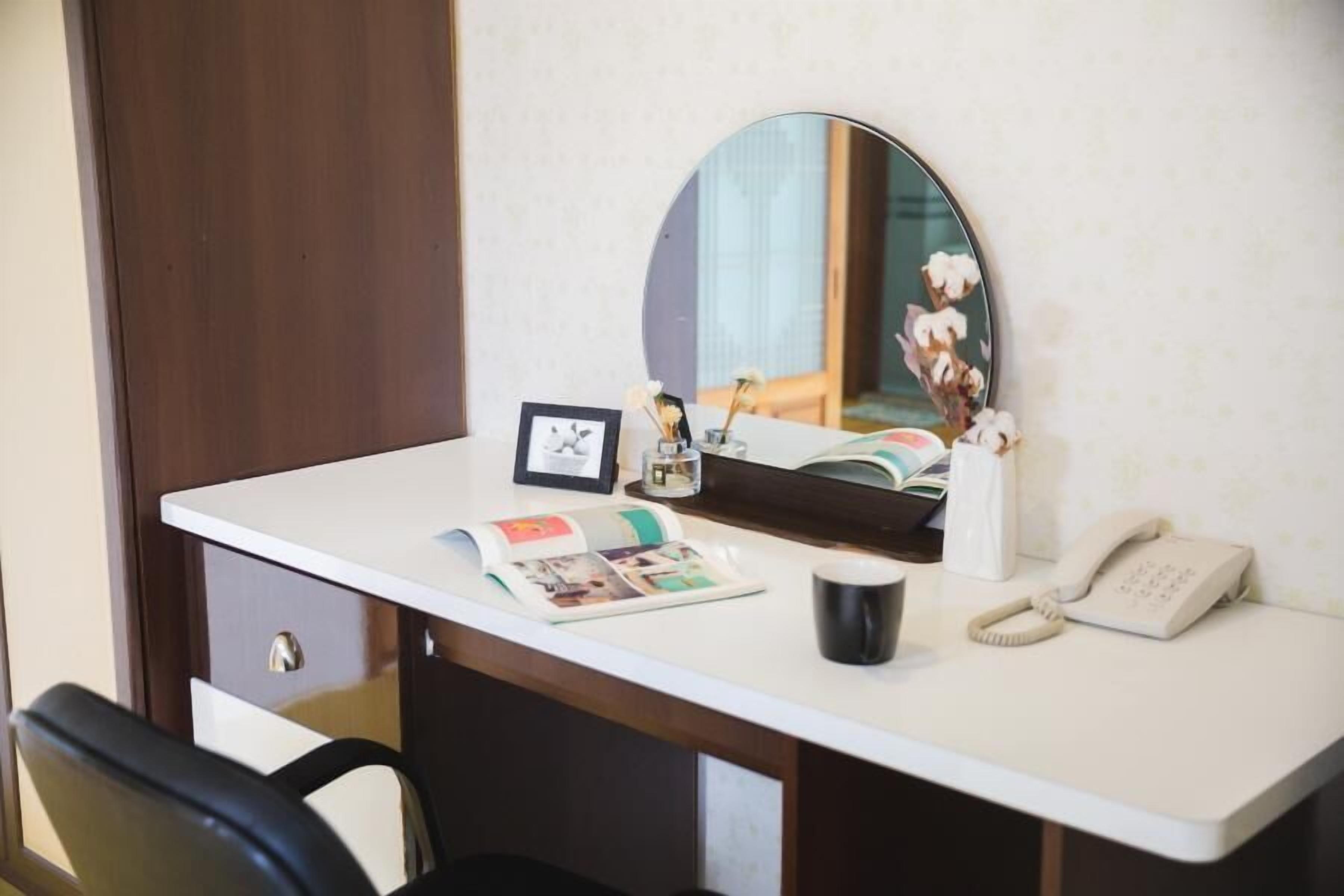 A hotel desk with a mirror, a framed photo, a book, a phone, and a cup.