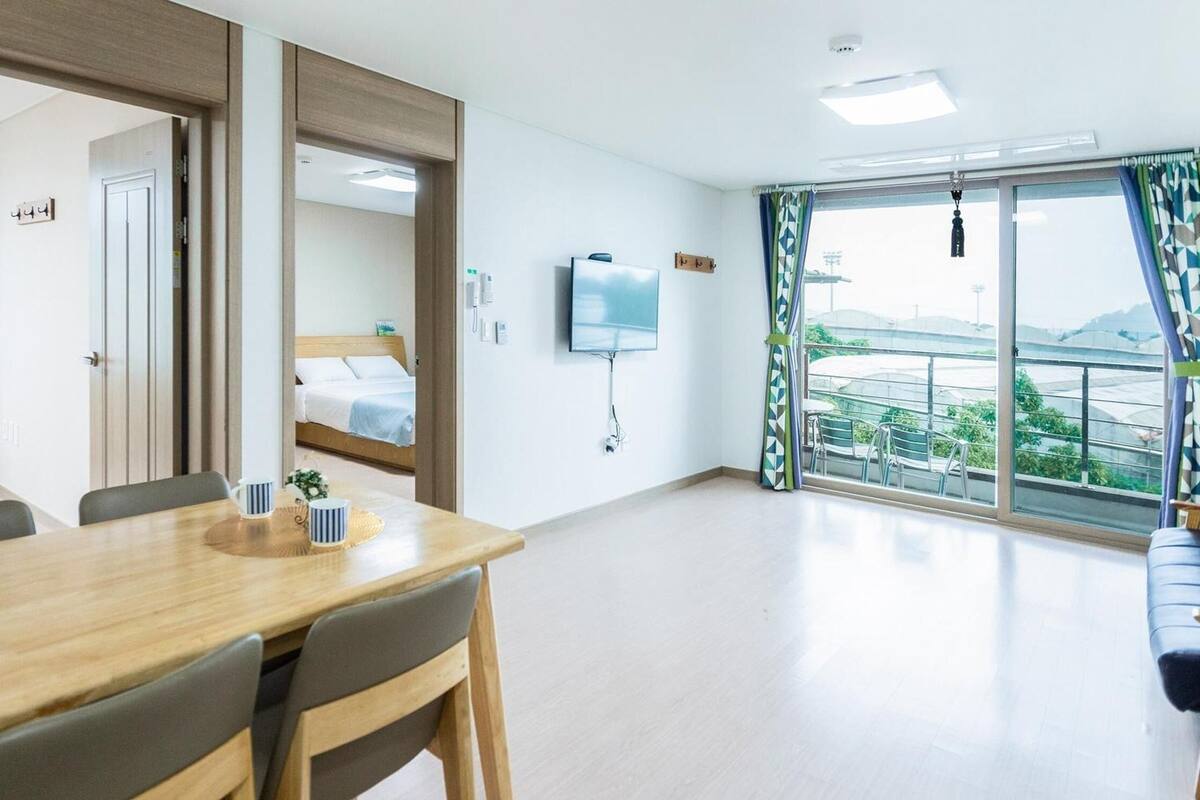 Een hotelkamer met een houten eettafel, stoelen, een bed met een wit hoofdbord, een aan de muur bevestigde televisie en een balkon met uitzicht op het omliggende landschap.