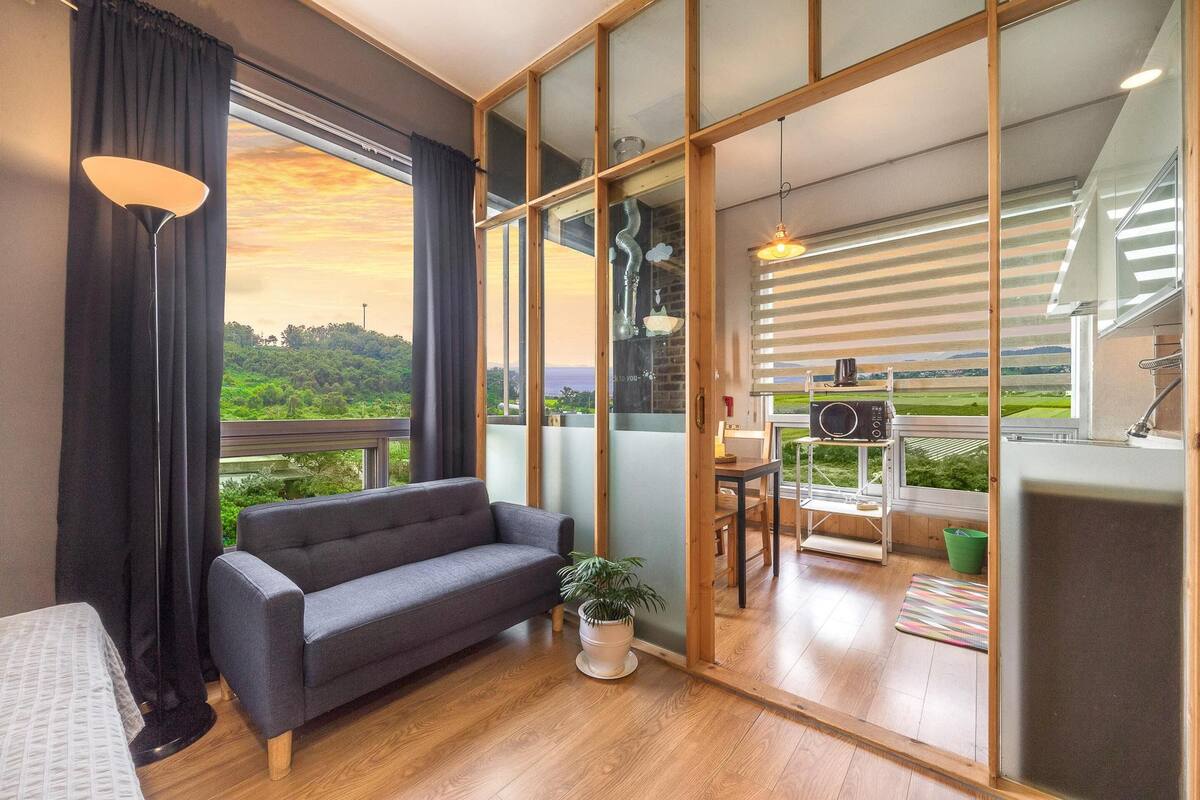 Een moderne kamer met een grijze bank, een tafeltje met een plant en uitzicht op een groen landschap door grote ramen.