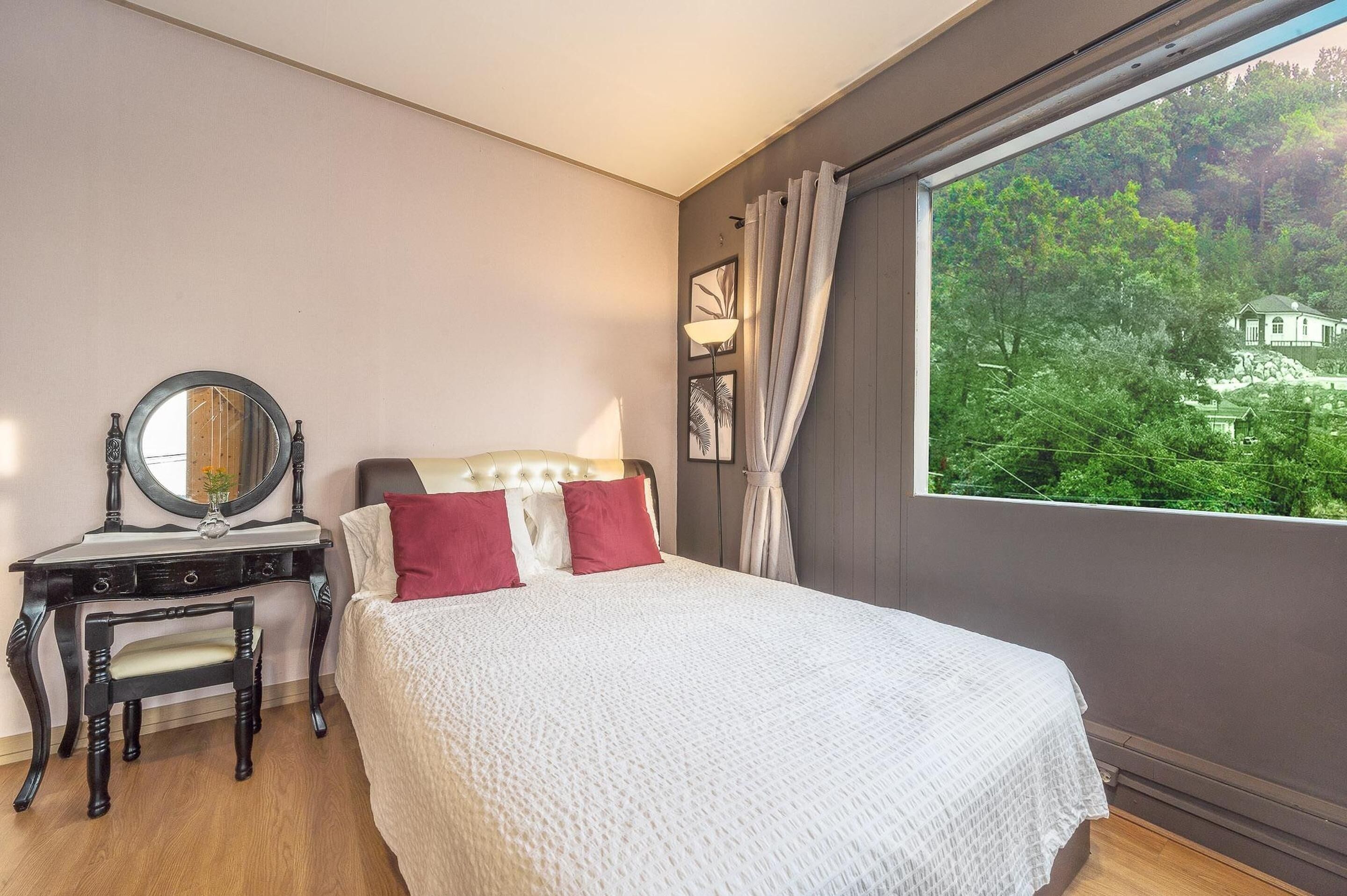 Ein Schlafzimmer mit Bett, Schminktisch mit Spiegel und Blick auf ein Haus durch ein Fenster.