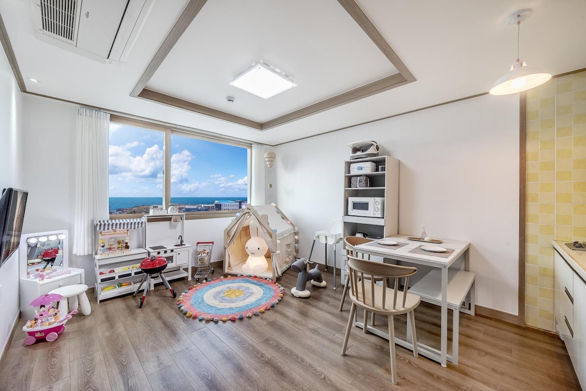 Una habitación infantil con un ventanal grande, una casita de juegos, un tocador con espejo y una pequeña mesa de comedor.