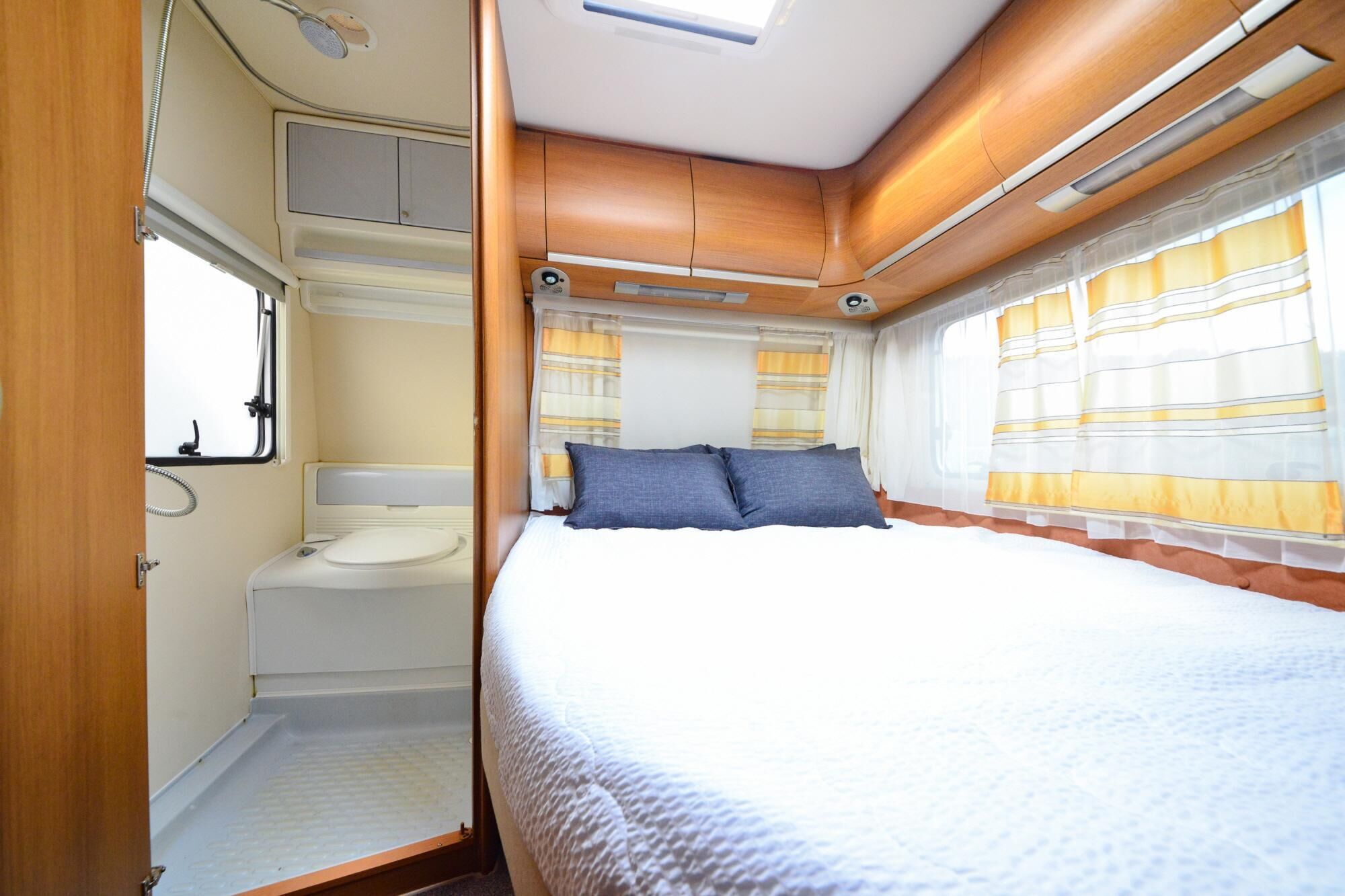 A compact RV interior with a bed, a small bathroom, and a window with curtains.