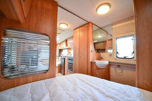 Interior compacto de autocaravana con cama, kitchenette, baño con lavabo y ventana.