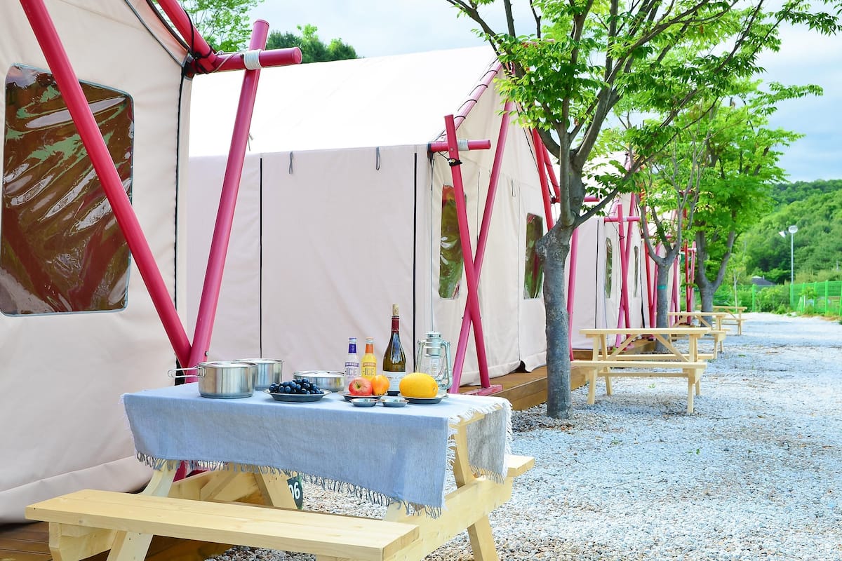 A row of white tents with pink poles, a table with a blue tablecloth, and a wooden bench.
