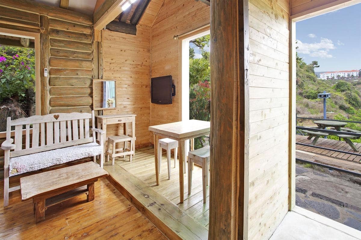 Une cabane en bois avec un banc, une table et des chaises à l’intérieur, et une vue sur un espace de pique-nique à l’extérieur.